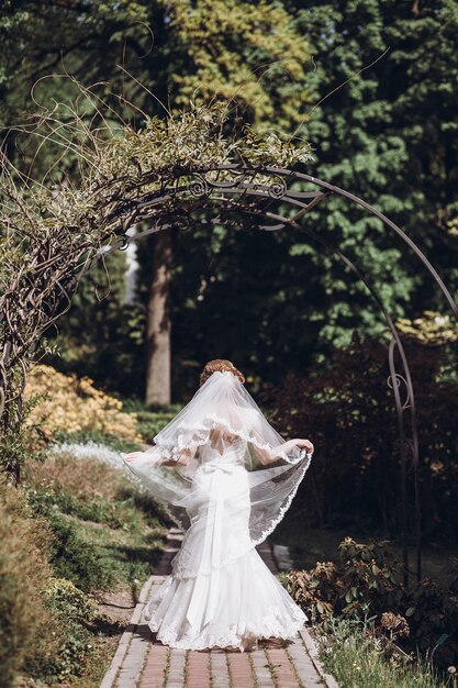 Belle mariée magnifique avec voile posant et s'amusant dans le parc de printemps ensoleillé femme heureuse debout dans le jardin vert heureux mariage moments concept de fugue