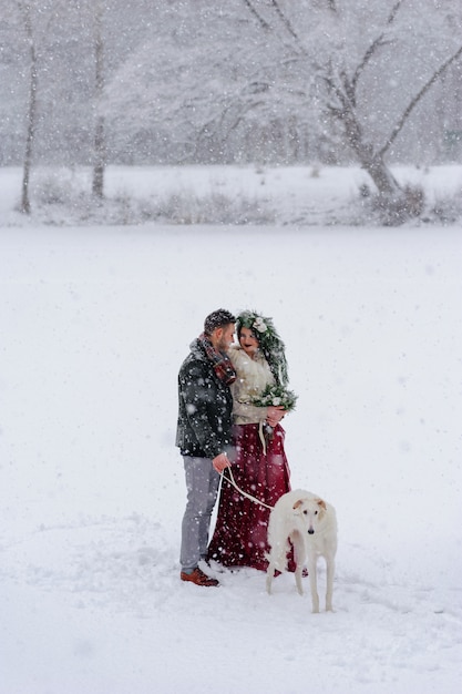 Belle mariée et le marié avec un chien blanc sont debout sur le fond d'une forêt enneigée
