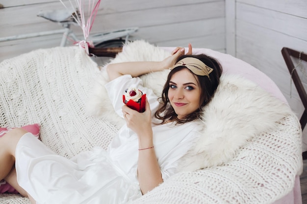 Belle mariée en masque de sommeil et robe avec cupcake sur un canapé confortable