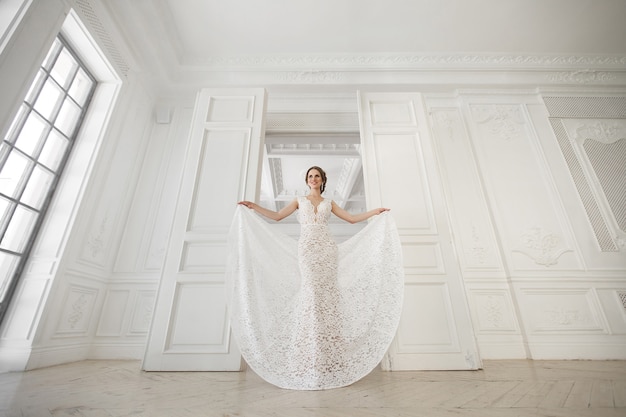 Belle mariée posant en robe de mariée dans un studio photo blanc. Miroir. Canapé. Bouquet. La porte. Lustre.