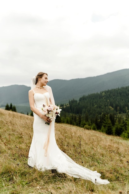 Une belle mariée en robe blanche et avec un bouquet de mariage de fleurs séchées est dans les montagnes d'automne parmi l'herbe jaune