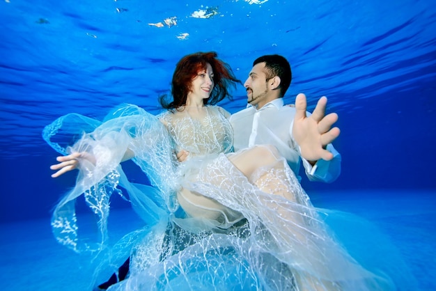 Une belle mariée vêtue d'une robe blanche comme neige entre les mains du marié nage sous l'eau dans la piscine