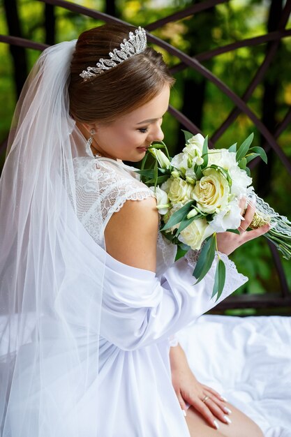Belle mariée en voile et manteau blanc est assise sur une couverture avec un bouquet de mariage dans les mains