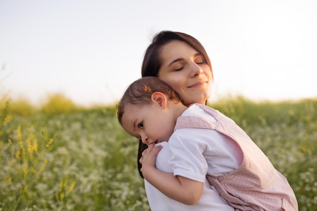 Belle maternité jeune jolie mère aux yeux fermés embrasse un bébé fille tout-petit sur les mains sur fond de nature de champ