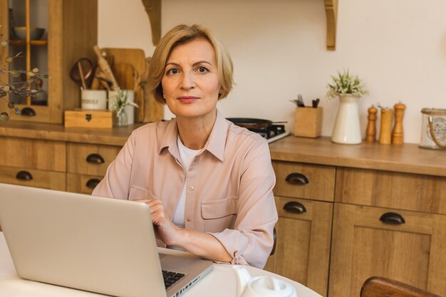 Belle matinée. Joyeuse femme âgée souriante debout dans la cuisine et utilisant son ordinateur portable tout en se reposant après le petit-déjeuner, pigiste travaillant à la maison.