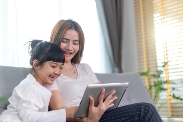 Belle mère asiatique avec sa fille assise sur le canapé et jouant à un jeu vidéo sur une tablette tout en passant du temps ensemble à la maison