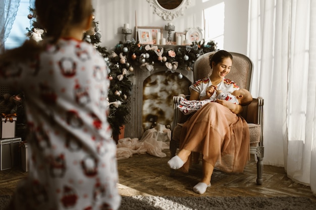 Belle mère est assise dans le fauteuil avec son petit bébé à côté de la cheminée et de l'arbre du Nouvel An avec des cadeaux dans la pièce confortable et lumineuse et sa petite fille court. .