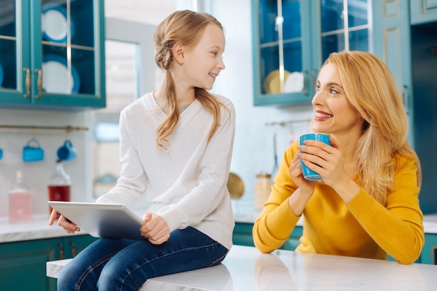 Belle mère mince blonde exubérante souriant et tenant une tasse de thé et sa fille à l'aide d'une tablette alors qu'il était assis sur la table