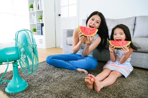 belle mère avec sa fille mangeant de la pastèque profitant du vent frais du ventilateur électrique à la maison en été et regardant la caméra en souriant.