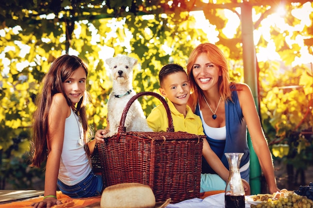 Belle mère souriante et ses enfants mignons avec un chien en train de pique-niquer dans un vignoble. Regarder la caméra.