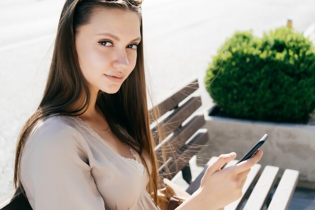 Belle et mignonne fille assise sur un banc et grimpant dans le téléphone et regardant la caméra