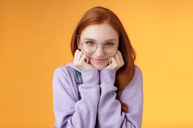 Belle mignonne rousse douce fille idiote geek étudiant universitaire portant des lunettes main maigre souriant tendrement regarder affection adorer écouter des confessions sensuelles petit ami, debout fond orange.