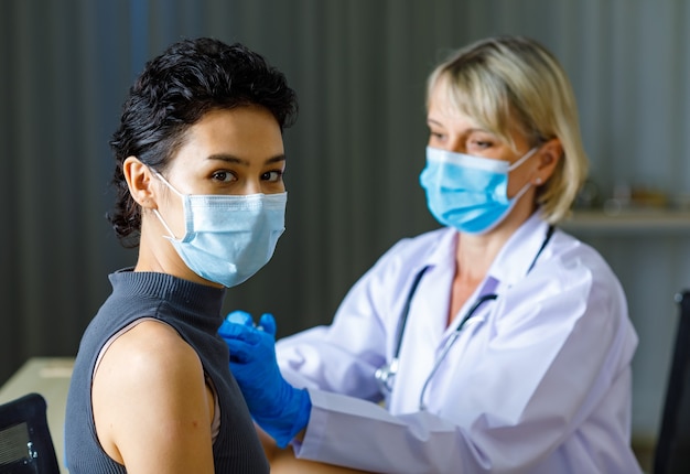 Photo une belle patiente aux cheveux noirs et courts porte un masque facial assis regarde la caméra tandis qu'un médecin caucasien en blouse blanche avec stéthoscope injecte le vaccin dans son épaule en arrière-plan flou.