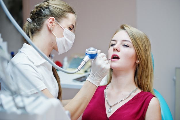 Belle patiente vêtue d’une robe rouge violette, allongée sur le fauteuil dentaire, pendant que la dentiste traite ses dents avec des instruments spéciaux.