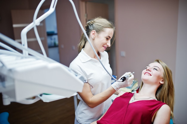 Belle patiente vêtue d’une robe rouge violette, allongée sur le fauteuil dentaire, pendant que la dentiste traite ses dents avec des instruments spéciaux.