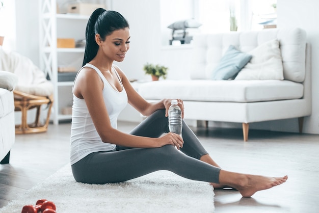 Belle perfection ! Jolie jeune femme tenant une bouteille et regardant ailleurs alors qu'elle était assise sur le tapis à la maison