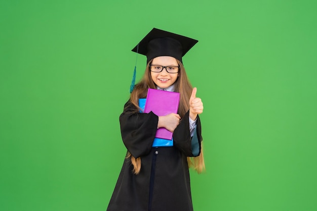 Une belle petite écolière dans un bonnet de graduation et une robe de cérémonie, tenant des livres sur un fond vert isolé. et montrer un geste de la main tout est super.