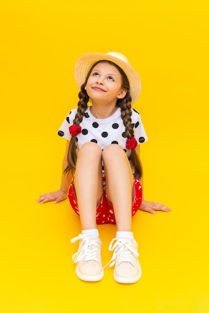 Une belle petite écolière dans un chapeau de paille lève les yeux et sourit largement Une adolescente est assise dans des vêtements d'été sur un fond isolé jaune