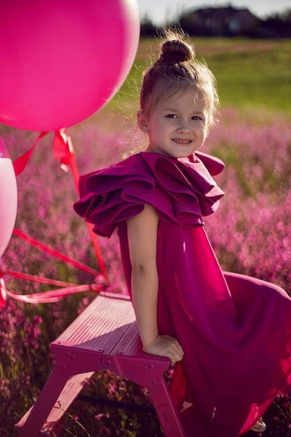 Belle petite fille de 5 ans est assise sur un escabeau rose dans une robe rose dans un champ de fleurs roses en été au coucher du soleil