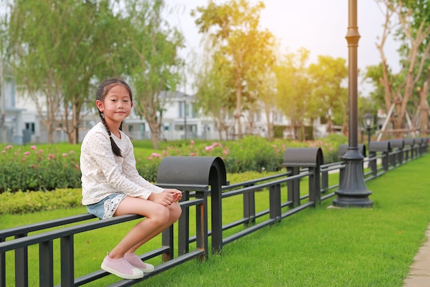 Belle petite fille asiatique assise dans le jardin sur une clôture au parc public en plein air