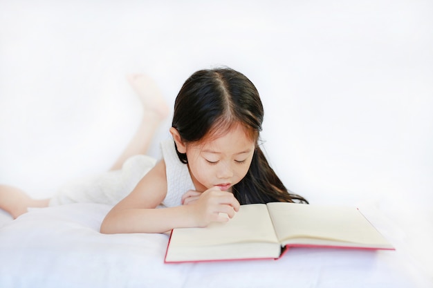 Belle petite fille asiatique enfant lisant un livre relié, couché avec oreiller sur fond blanc.