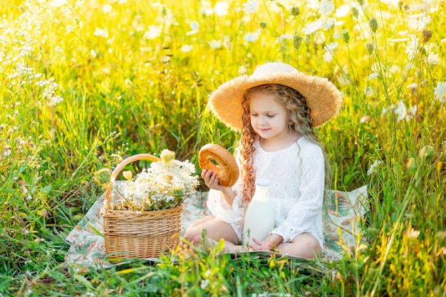 Belle petite fille assise dans un chapeau de paille dans un champ jaune avec des fleurs sauvages avec une bouteille de lait et un bagel
