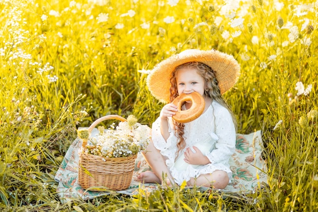 Belle petite fille assise dans un chapeau de paille dans un champ jaune avec des fleurs sauvages avec une bouteille de lait et un bagel