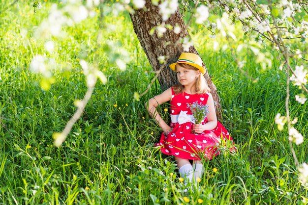 Belle petite fille assise sous un arbre avec un bouquet de fleurs sauvages dans ses mains