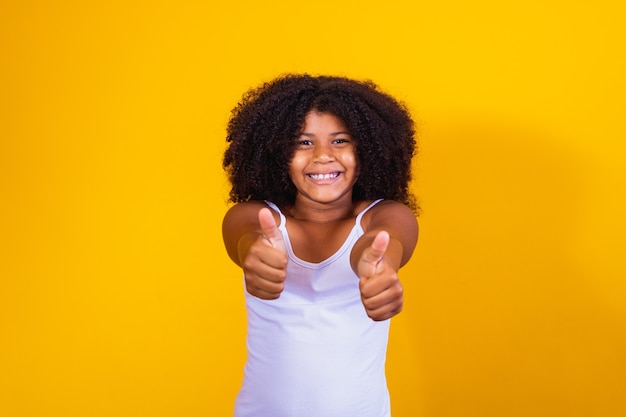 Belle petite fille aux cheveux bouclés utilisant ses mains signe ok. Fond jaune. Fille afro ok pouce vers le haut