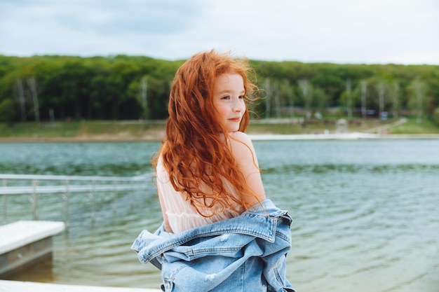 Une belle petite fille aux cheveux longs rouges se dresse sur la rive du portrait romantique du lac