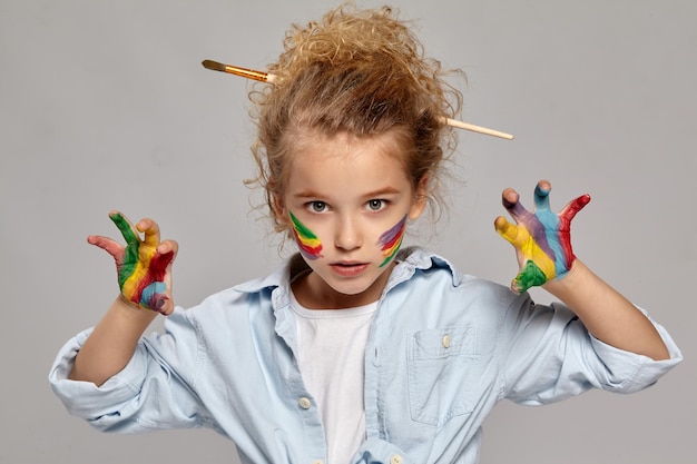 Belle petite fille ayant une brosse dans ses cheveux blonds bouclés chics, vêtue d'une chemise bleue et d'un t-shirt blanc, avec des doigts peints, agit comme si elle faisait peur à quelqu'un, sur fond gris.