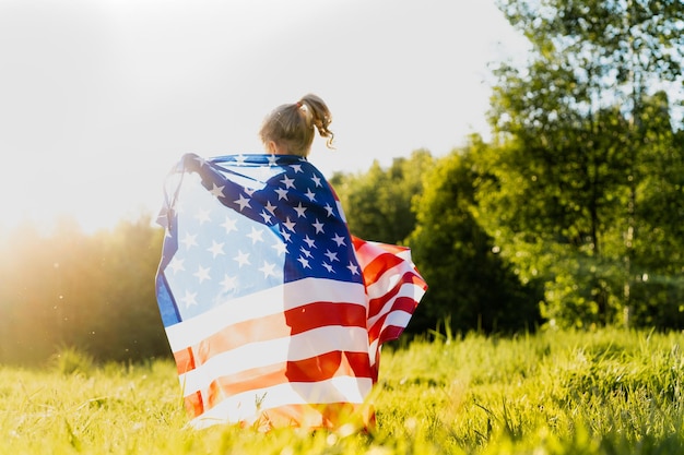 belle petite fille blonde avec le drapeau américain dans la nature au soleil.