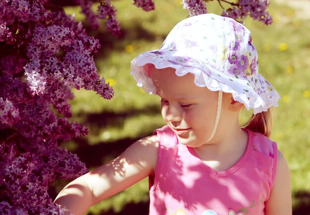 Belle petite fille blonde avec un panama sur la tête jouant dans un jardin de printemps avec un buisson en fleurs de lilas violet