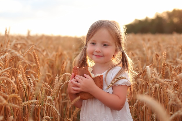 une belle petite fille blonde en vêtements blancs se dresse dans un champ de blé avec du pain