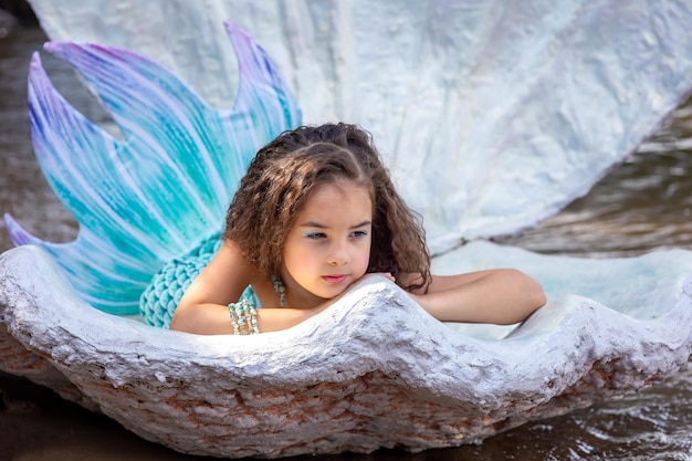 Une belle petite fille brune dans un costume de sirène turquoise se trouve dans un grand coquillage en plein air en détournant les yeux Close up Copy space