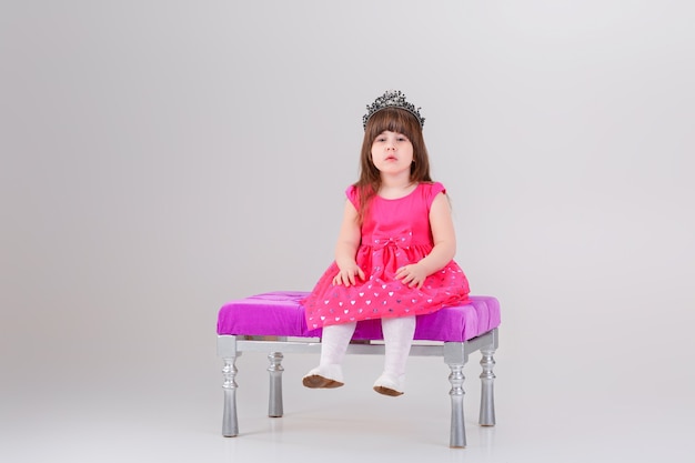 Belle petite fille brune en robe de princesse rose avec couronne assise sur une chaise rose sur fond gris. bébé mignon