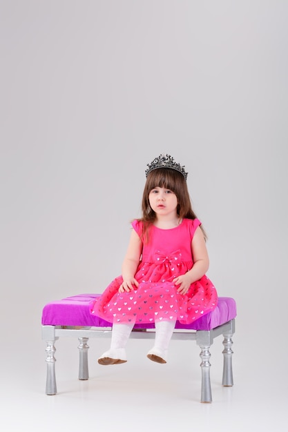 Belle petite fille brune en robe de princesse rose avec couronne assise sur une chaise rose sur fond gris. bébé mignon