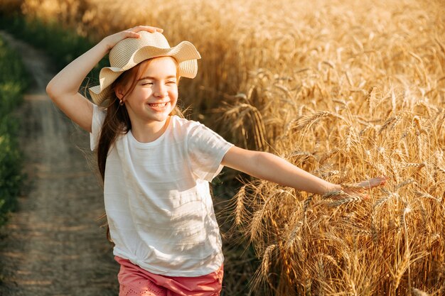 Belle petite fille avec un chapeau sur la tête et un sourire attrayant court sur la route de campagne près du champ...