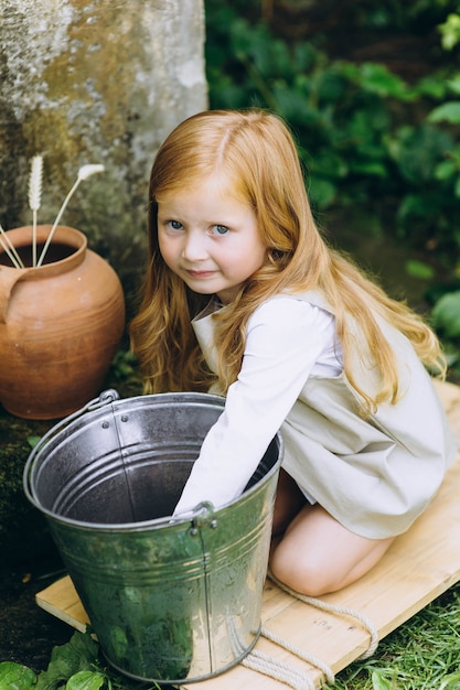 Belle petite fille avec une cruche et un seau près du puits à côté des arbres et des herbes