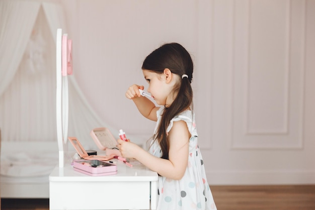 Une belle petite fille dans le miroir se lissant une petite fille est assise à une table pour enfants et porte des cosmétiques pour enfants