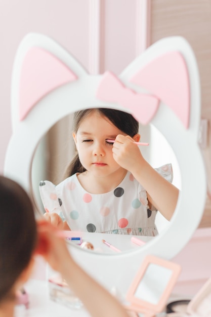 Une belle petite fille dans le miroir se lissant une petite fille est assise à une table pour enfants et porte des cosmétiques pour enfants