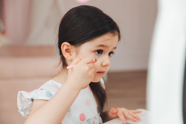 Une belle petite fille dans le miroir se lissant une petite fille est assise à une table pour enfants et porte des cosmétiques pour enfants
