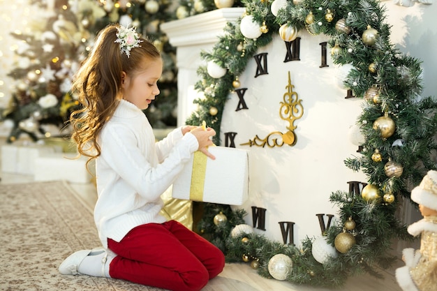 Belle petite fille dans un pull blanc se trouve près de l'arbre de Noël avec un cadeau à la main