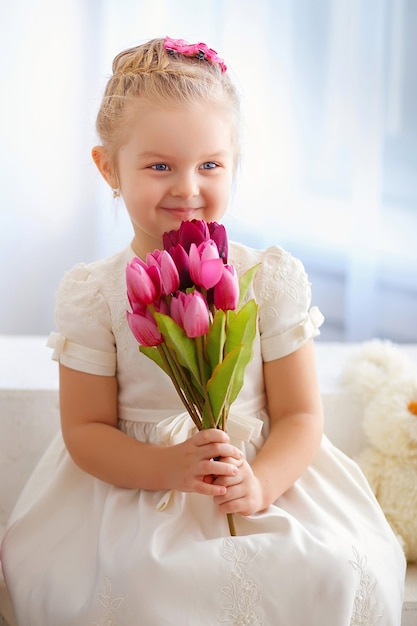 Belle petite fille dans une robe blanche assise sur un rebord de fenêtre avec un bouquet de tulipes roses