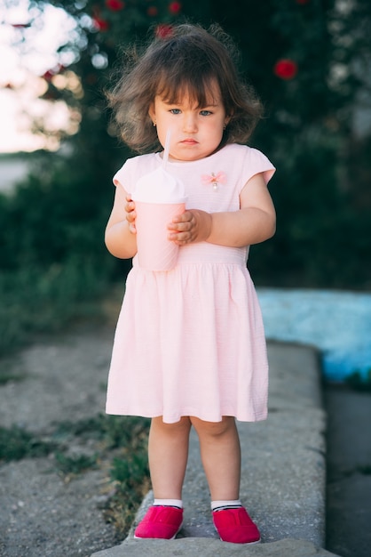 Une belle petite fille dans une robe rose sur fond de plantes boit un verre dans un verre à cocktail mignon