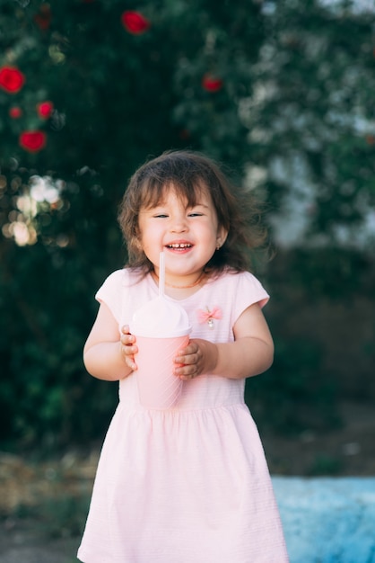 Une belle petite fille dans une robe rose se dresse sur un fond de plantes et tient un cocktail rose