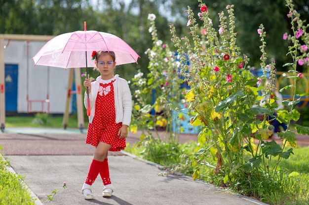 Une belle petite fille dans une robe rouge et un chemisier blanc avec un parapluie rose