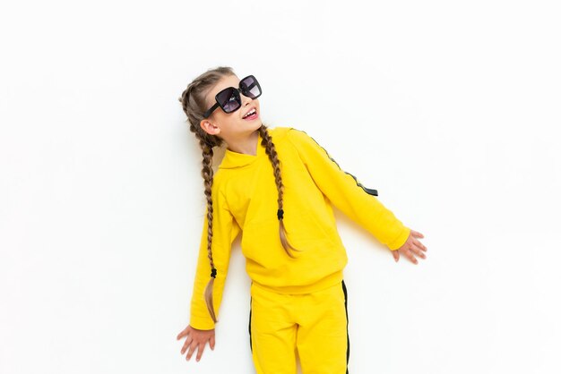 Une belle petite fille dans un survêtement jaune avec des nattes sur la tête et des lunettes de soleil sur un fond blanc isolé