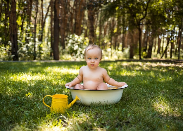 Une belle petite fille est assise dans une baignoire avec de l'eau et un arrosoir et regarde la caméra sur l'herbe verte