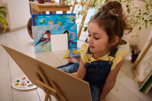 Belle petite fille européenne appréciant l'apprentissage de la peinture assise sur le sol sur un chevalet en bois sur fond d'images peintes d'enfants joyeux et colorés Créativité et éducation de la classe d'art